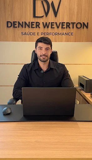 Denner Weverton sentado à mesa do consultório, com o painel de madeira da marca ao fundo